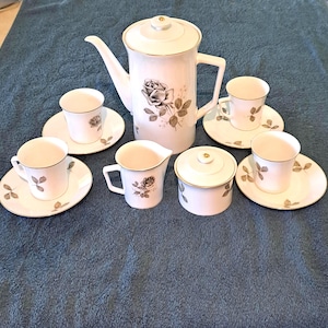 May include: A white porcelain tea set with a floral rose design. The set includes a teapot, creamer, sugar bowl, and four teacups with saucers. The rims and accents are gold. The set is arranged on a blue surface.