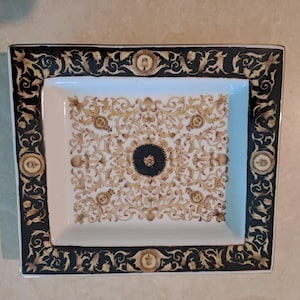 May include: Rectangular decorative tray with a white interior and a black border. The border features gold and black ornate scrollwork and medallions. The tray's interior has a gold and white floral design with a black centre.