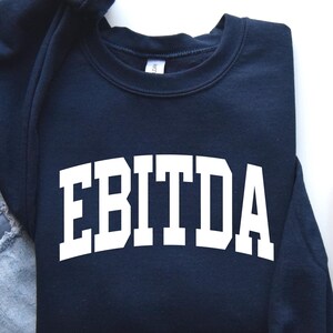 May include: A navy blue sweatshirt with the word "EBITDA" in bold white letters. The sweatshirt is laid flat next to a pair of light blue jeans, a brown mug, and gray sneakers.