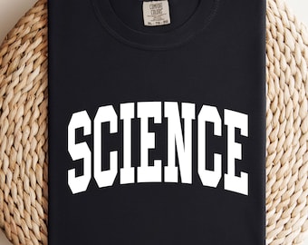 Pode incluir: Uma camiseta preta com a palavra "SCIENCE" impressa em letras brancas grandes. A camiseta está dobrada e repousa sobre um sousplat redondo e tecido bege. A camiseta tem gola redonda e mangas curtas.