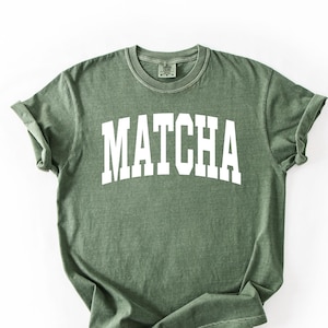 May include: Olive green t-shirt with the word "MATCHA" printed in white block letters. The shirt has a crew neck and rolled-up sleeves. The fabric appears soft and slightly faded, giving it a vintage look.