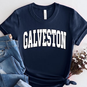 May include: Navy blue t-shirt with the word "GALVESTON" in white, block letters. The shirt is paired with a pair of blue jeans. A small bouquet of white flowers is placed next to the shirt.
