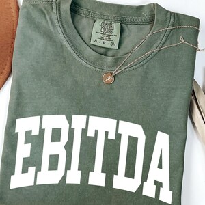 May include: An olive green t-shirt with the word "EBITDA" printed in large white block letters. A gold necklace with a pendant rests on the shirt. The shirt is a Comfort Colors brand, size S-P-CH.