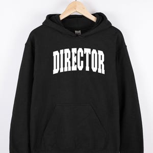 May include: Black hooded sweatshirt with the word "DIRECTOR" printed in large, white, block letters across the chest. The sweatshirt has a front pocket and a drawstring hood. The garment is hanging on a wooden hanger.