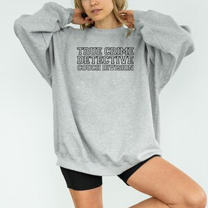 May include: A light gray sweatshirt with the text "TRUE CRIME DETECTIVE COUCH DIVISION" in black block letters. The sweatshirt has long sleeves and a crew neck. The overall style is casual and comfortable.