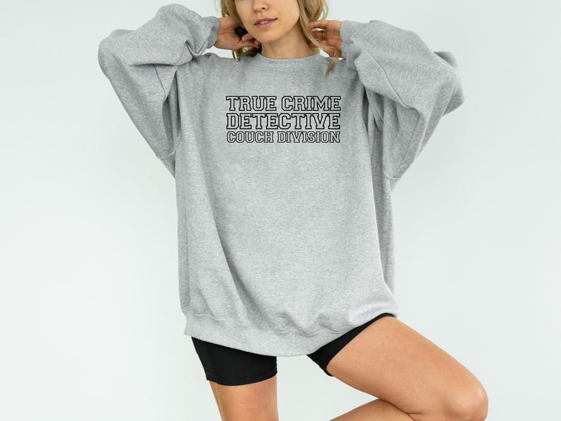May include: A light gray sweatshirt with the text "TRUE CRIME DETECTIVE COUCH DIVISION" in black block letters. The sweatshirt has long sleeves and a crew neck. The overall style is casual and comfortable.