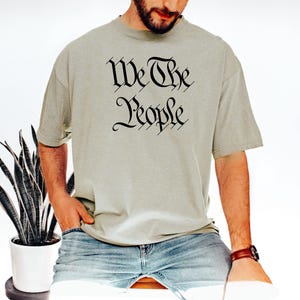 K&ouml;nnte beinhalten: Hellgr&uuml;nes T-Shirt mit dem Text "We The People" in schwarzer gotischer Schrift. Das Shirt wird mit einer blauen Jeans kombiniert. Im Hintergrund ist eine Topfpflanze zu sehen.