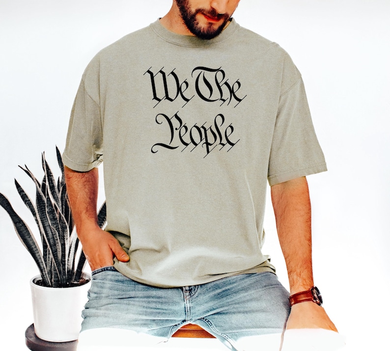 K&ouml;nnte beinhalten: Hellgr&uuml;nes T-Shirt mit dem Text "We The People" in schwarzer gotischer Schrift. Das Shirt wird mit einer blauen Jeans kombiniert. Im Hintergrund ist eine Topfpflanze zu sehen.