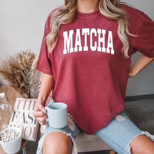 K&ouml;nnte beinhalten: Ein weinrotes T-Shirt mit dem Wort "MATCHA" in gro&szlig;en wei&szlig;en Blockbuchstaben. Die Person tr&auml;gt eine hellblaue zerrissene Jeans und h&auml;lt eine hellblaue Tasse. Im Hintergrund ein Tisch mit einem Buch und einer Schale mit wei&szlig;en Steinen.