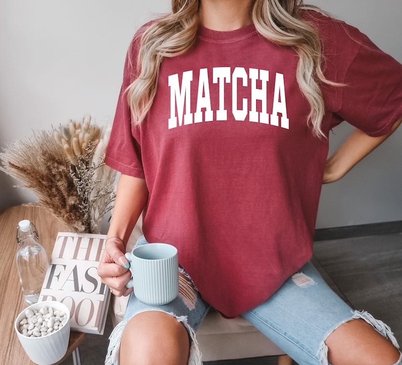 K&ouml;nnte beinhalten: Ein weinrotes T-Shirt mit dem Wort "MATCHA" in gro&szlig;en wei&szlig;en Blockbuchstaben. Die Person tr&auml;gt eine hellblaue zerrissene Jeans und h&auml;lt eine hellblaue Tasse. Im Hintergrund ein Tisch mit einem Buch und einer Schale mit wei&szlig;en Steinen.