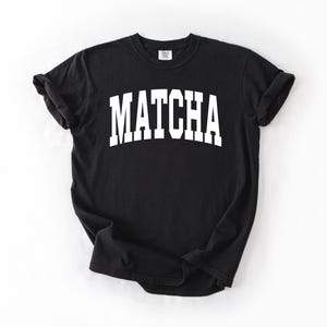 K&ouml;nnte beinhalten: Schwarzes T-Shirt mit dem Wort "MATCHA" in gro&szlig;en, wei&szlig;en Blockbuchstaben auf der Vorderseite. Das Shirt hat kurze &Auml;rmel und einen Rundhalsausschnitt.