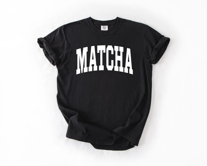 K&ouml;nnte beinhalten: Schwarzes T-Shirt mit dem Wort "MATCHA" in gro&szlig;en, wei&szlig;en Blockbuchstaben auf der Vorderseite. Das Shirt hat kurze &Auml;rmel und einen Rundhalsausschnitt.