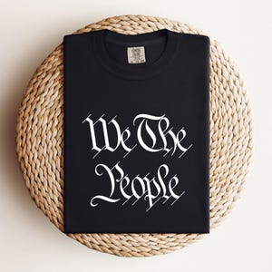 K&ouml;nnte beinhalten: Ein schwarzes T-Shirt mit dem wei&szlig;en Schriftzug "We The People" in einer verzierten Schriftart. Das T-Shirt ist gefaltet und liegt auf einem runden, gewebten, beigefarbenen Platzdeckchen. Rundhalsausschnitt, kurze &Auml;rmel.