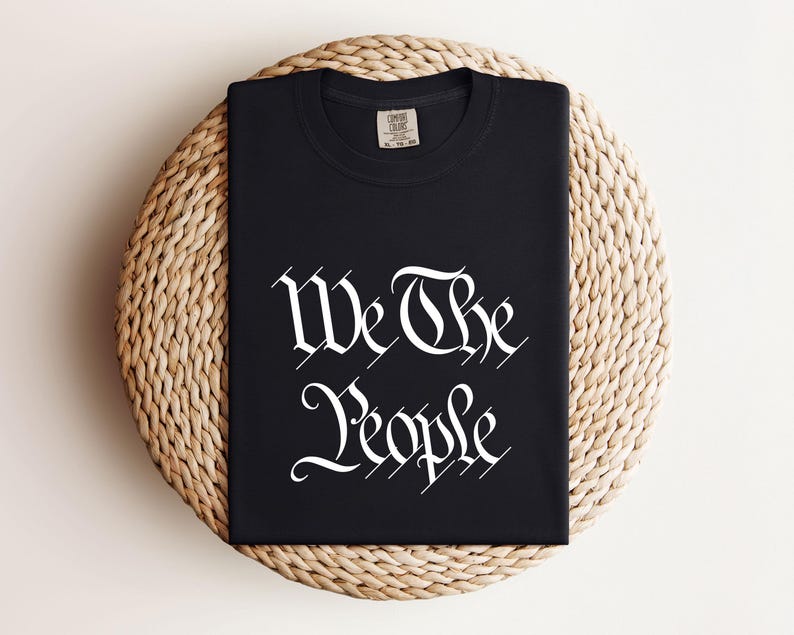 K&ouml;nnte beinhalten: Ein schwarzes T-Shirt mit dem wei&szlig;en Schriftzug "We The People" in einer verzierten Schriftart. Das T-Shirt ist gefaltet und liegt auf einem runden, gewebten, beigefarbenen Platzdeckchen. Rundhalsausschnitt, kurze &Auml;rmel.