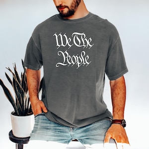 K&ouml;nnte beinhalten: Dunkelgraues T-Shirt mit dem wei&szlig;en Text "We The People" in einer eleganten Schriftart. Das T-Shirt hat einen Rundhalsausschnitt und scheint aus einem weichen Material zu sein. Das Model tr&auml;gt eine Jeans und eine Armbanduhr.