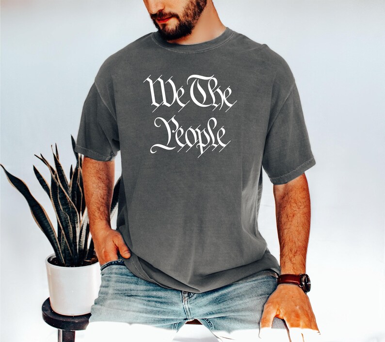 K&ouml;nnte beinhalten: Dunkelgraues T-Shirt mit dem wei&szlig;en Text "We The People" in einer eleganten Schriftart. Das T-Shirt hat einen Rundhalsausschnitt und scheint aus einem weichen Material zu sein. Das Model tr&auml;gt eine Jeans und eine Armbanduhr.