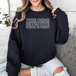 May include: A black sweatshirt with the text "TRUE CRIME DETECTIVE COUCH DIVISION" in white block letters. The sweatshirt is a crew neck style and appears to be made of a soft material.