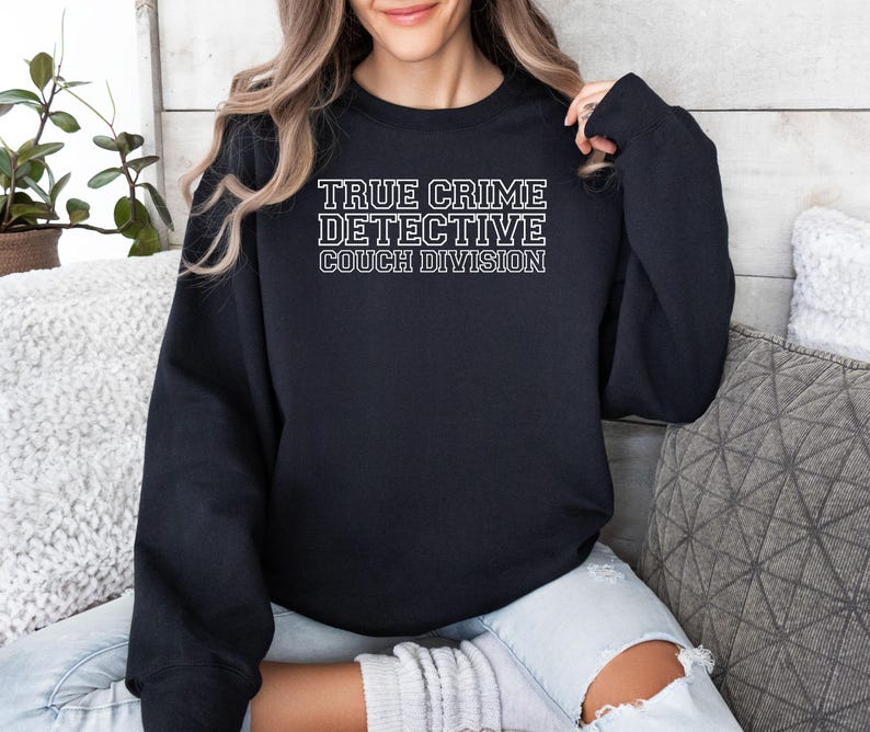 May include: A black sweatshirt with the text "TRUE CRIME DETECTIVE COUCH DIVISION" in white block letters. The sweatshirt is a crew neck style and appears to be made of a soft material.
