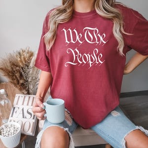 K&ouml;nnte beinhalten: Ein weinrotes T-Shirt mit dem wei&szlig;en Schriftzug "We The People" in stilisierten Buchstaben. Das Shirt wird mit einer blauen Jeans getragen. Eine hellblaue Tasse wird in der Hand gehalten.