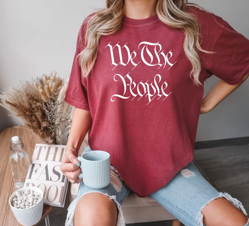 K&ouml;nnte beinhalten: Ein weinrotes T-Shirt mit dem wei&szlig;en Schriftzug "We The People" in stilisierten Buchstaben. Das Shirt wird mit einer blauen Jeans getragen. Eine hellblaue Tasse wird in der Hand gehalten.