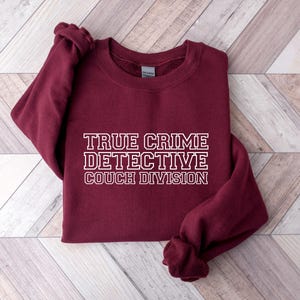 May include: A maroon sweatshirt with the text "TRUE CRIME DETECTIVE COUCH DIVISION" in white block letters. The sweatshirt is folded, with the sleeves and bottom hem visible. The background is a light wood grain pattern.