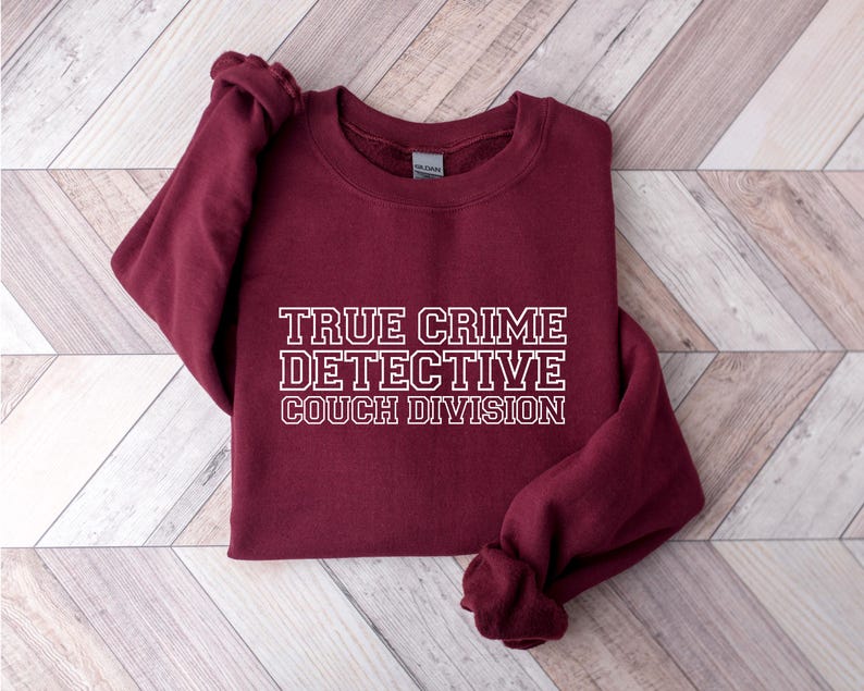 May include: A maroon sweatshirt with the text "TRUE CRIME DETECTIVE COUCH DIVISION" in white block letters. The sweatshirt is folded, with the sleeves and bottom hem visible. The background is a light wood grain pattern.