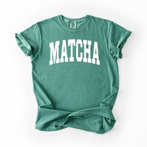 K&ouml;nnte beinhalten: Ein teal-farbenes T-Shirt mit dem Wort "MATCHA" in gro&szlig;en, wei&szlig;en Blockbuchstaben auf der Brust. Das Shirt hat einen Rundhalsausschnitt und kurze &Auml;rmel, die hochgekrempelt sind. Der Stoff wirkt weich und bequem.