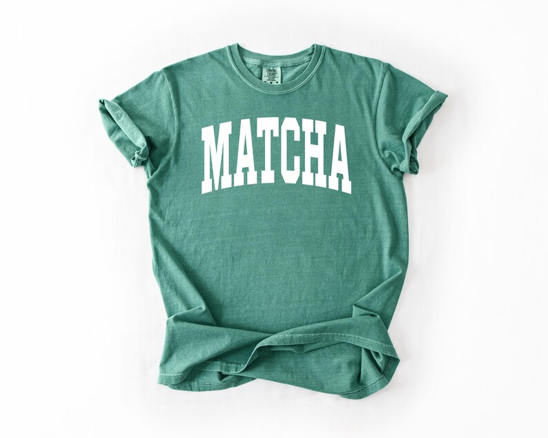 K&ouml;nnte beinhalten: Ein teal-farbenes T-Shirt mit dem Wort "MATCHA" in gro&szlig;en, wei&szlig;en Blockbuchstaben auf der Brust. Das Shirt hat einen Rundhalsausschnitt und kurze &Auml;rmel, die hochgekrempelt sind. Der Stoff wirkt weich und bequem.