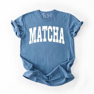 K&ouml;nnte beinhalten: Ein blaues T-Shirt mit dem Wort "MATCHA" in gro&szlig;en, wei&szlig;en Blockbuchstaben aufgedruckt. Das Shirt hat kurze &Auml;rmel und einen Rundhalsausschnitt. Der Stoff wirkt weich und leicht verblichen.