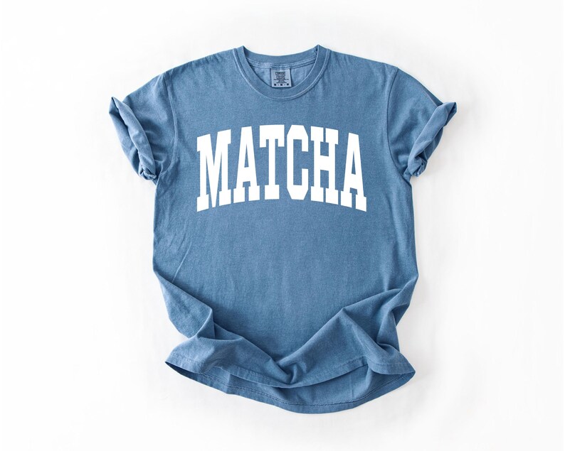 K&ouml;nnte beinhalten: Ein blaues T-Shirt mit dem Wort "MATCHA" in gro&szlig;en, wei&szlig;en Blockbuchstaben aufgedruckt. Das Shirt hat kurze &Auml;rmel und einen Rundhalsausschnitt. Der Stoff wirkt weich und leicht verblichen.