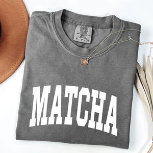K&ouml;nnte beinhalten: Ein graues T-Shirt mit dem Wort "MATCHA" in gro&szlig;en wei&szlig;en Blockbuchstaben. Eine goldene Halskette mit einem kleinen Anh&auml;nger liegt auf dem Shirt. Ein brauner Hut und eine getrocknete Pflanze sind im Hintergrund.