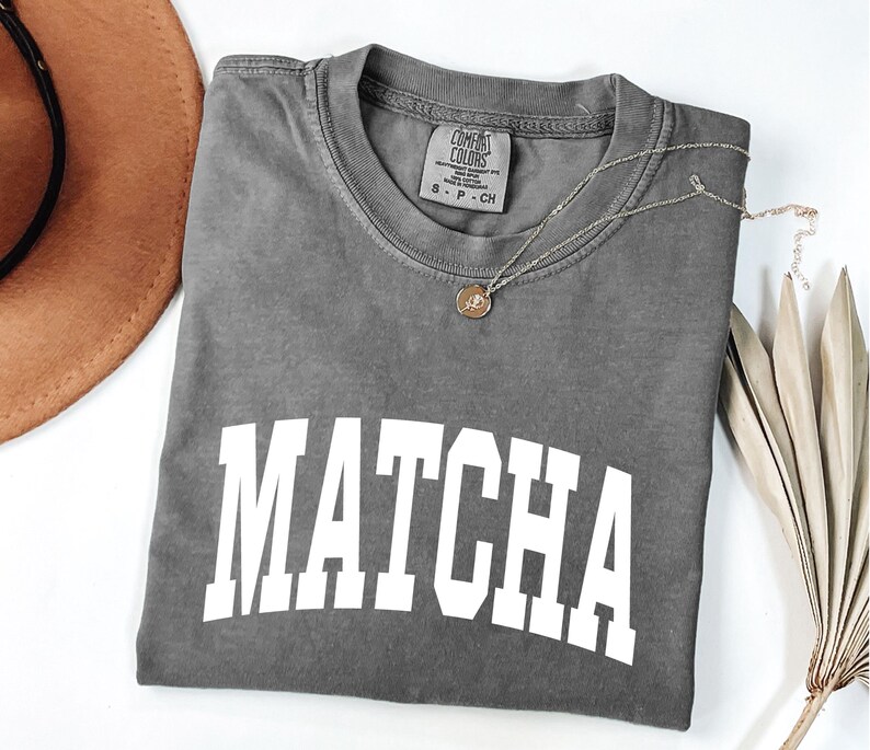 K&ouml;nnte beinhalten: Ein graues T-Shirt mit dem Wort "MATCHA" in gro&szlig;en wei&szlig;en Blockbuchstaben. Eine goldene Halskette mit einem kleinen Anh&auml;nger liegt auf dem Shirt. Ein brauner Hut und eine getrocknete Pflanze sind im Hintergrund.