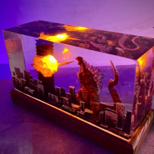 Peut inclure: Un bloc de r&eacute;sine transparent contenant une ville miniature et une figurine rouge de Godzilla. La ville est &eacute;clair&eacute;e par derri&egrave;re, cr&eacute;ant un effet lumineux. La figurine de Godzilla est en position de rugissement, la bouche ouverte et les bras tendus.