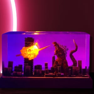 Peut inclure: Un bloc de r&eacute;sine transparent avec une ville miniature et une figurine de Godzilla. La figurine est rouge et noire et se tient sur un b&acirc;timent. Il y a une explosion jaune derri&egrave;re la figurine. Le bloc est &eacute;clair&eacute; de l'int&eacute;rieur par des lumi&egrave;res violettes et jaunes.