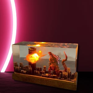 Peut inclure: Un bloc de r&eacute;sine transparent contenant une ville miniature et une figurine de Godzilla. La ville est &eacute;clair&eacute;e de l'int&eacute;rieur, cr&eacute;ant un effet lumineux. La base est en bois.