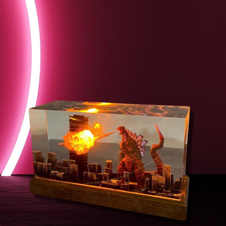Peut inclure: Un bloc de r&eacute;sine transparent contenant une ville miniature et une figurine de Godzilla. La ville est &eacute;clair&eacute;e de l'int&eacute;rieur, cr&eacute;ant un effet lumineux. La base est en bois.
