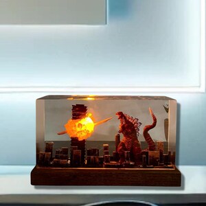 Peut inclure: Un bloc de r&eacute;sine transparent avec une ville miniature, une explosion orange incandescente et un monstre rouge ressemblant &agrave; Godzilla. La base est en bois fonc&eacute;.