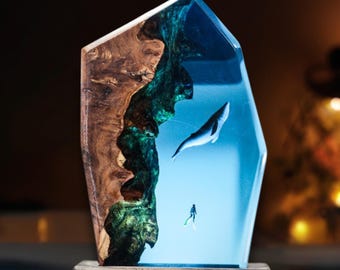 May include: A resin sculpture of a whale and a diver in a blue and green ocean scene. The sculpture is set on a wooden base.