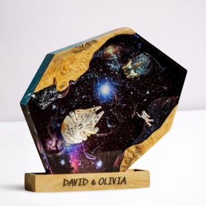 May include: A unique, geometric resin sculpture featuring a space scene with a spaceship, astronaut, and galaxy design. The sculpture has a wooden base with the names "DAVID & OLIVIA" engraved on it. The edges are natural wood.