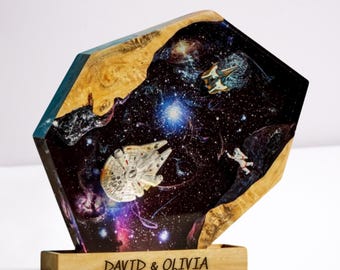 May include: A unique, geometric resin sculpture featuring a space scene with a spaceship, astronaut, and galaxy design. The sculpture has a wooden base with the names "DAVID & OLIVIA" engraved on it. The edges are natural wood.