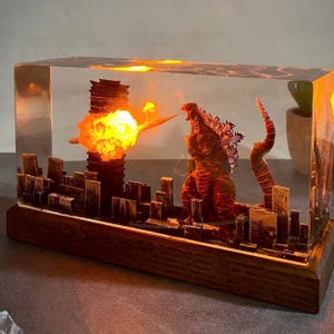 Peut inclure: Une sculpture en r&eacute;sine transparente de Godzilla d&eacute;truisant une ville. La sculpture est &eacute;clair&eacute;e de l'int&eacute;rieur, cr&eacute;ant un effet lumineux. La sculpture est sur une base en bois.