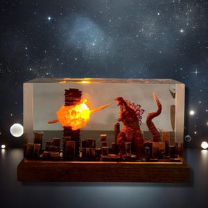 Peut inclure: Une sculpture en r&eacute;sine de Godzilla d&eacute;truisant une ville avec un faisceau d'&eacute;nergie. La sculpture est &eacute;clair&eacute;e de l'int&eacute;rieur, cr&eacute;ant un effet dramatique. La sculpture est pos&eacute;e sur une base en bois.