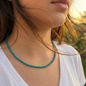 Puede incluir: Un collar de cuentas turquesas. El collar está compuesto por pequeñas cuentas redondas ensartadas para formar un delicado hilo. El collar se lleva sobre una camiseta blanca.