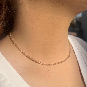 Puede incluir: Un delicado collar de cuentas doradas descansa sobre el cuello de una persona. El collar está compuesto por pequeñas cuentas espaciadas uniformemente, creando un efecto sutil y brillante. La persona lleva una camisa blanca y se ve un pequeño pendiente.