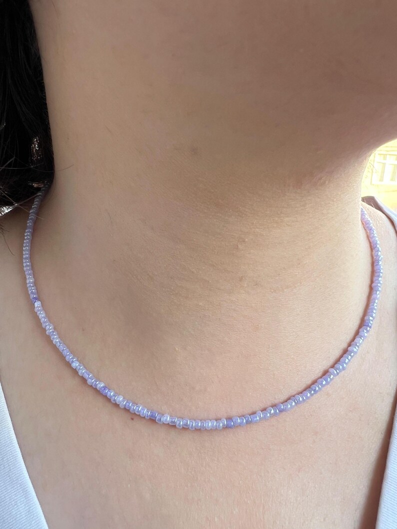 K&ouml;nnte beinhalten: Eine zarte Perlenkette in Lavendel- und Wei&szlig;t&ouml;nen. Die Halskette besteht aus kleinen, runden Perlen, die zu einem einfachen, eleganten Choker aufgereiht sind. Die Halskette wird um den Hals getragen.