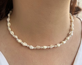 May include: A delicate necklace featuring small, cream-colored seashells and tiny, round pearls. The shells have natural patterns and textures, strung together with a fine, silver-colored chain. The necklace is a beach-inspired accessory.