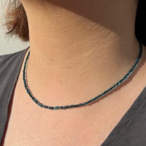 May include: A delicate beaded necklace in shades of teal and dark blue. The necklace is a choker style, resting closely against the neck. The beads appear to be small and evenly spaced, creating a subtle, shimmering effect. The necklace is worn against a gray top.
