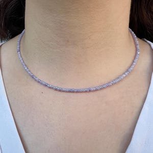 K&ouml;nnte beinhalten: Eine zarte, lavendelfarbene Perlenkette. Die Halskette besteht aus kleinen, runden Perlen, die ein subtiles und elegantes Accessoire bilden. Die Halskette wird um den Hals getragen und die Perlen sind pastellviolett.