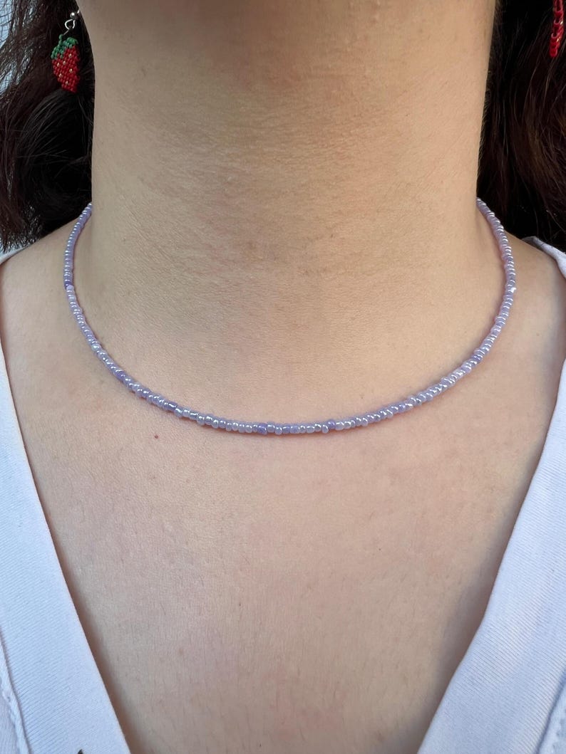 K&ouml;nnte beinhalten: Eine zarte, lavendelfarbene Perlenkette. Die Halskette besteht aus kleinen, runden Perlen, die ein subtiles und elegantes Accessoire bilden. Die Halskette wird um den Hals getragen und die Perlen sind pastellviolett.