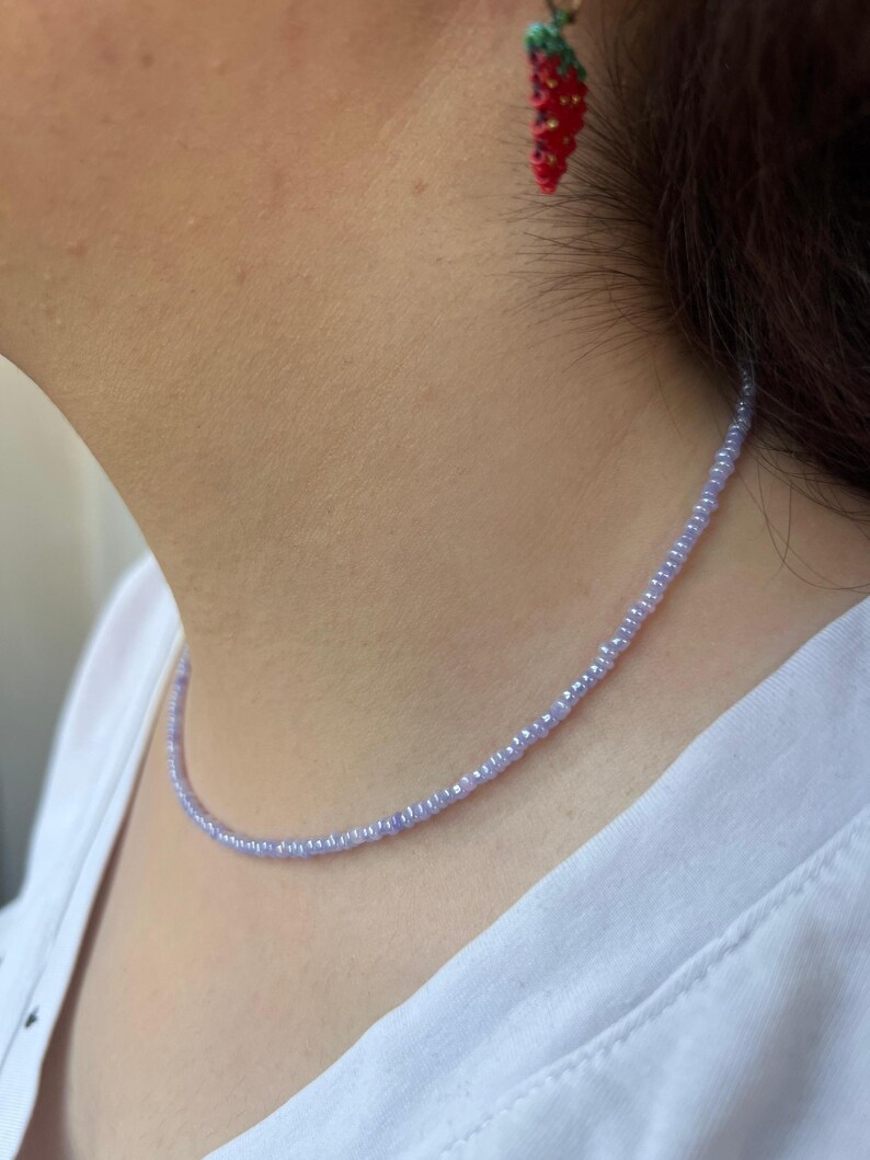 K&ouml;nnte beinhalten: Eine zarte, hellviolette Perlenkette aus kleinen, runden Perlen. Die Kette liegt am Hals. Im Hintergrund sind rote Erdbeerohrringe mit gr&uuml;nen Bl&auml;ttern zu sehen.
